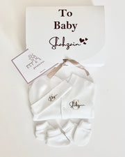 Personalized white baby gift set including an overall, socks, and a hat, presented in a gift box with a personalized message.