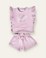 A set of lavender-colored kids' clothes, including a top with ruffles and shorts, with a personalized name 'Zaia' printed on the top.