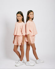 Two young girls wearing matching pink outfits standing side by side on a white background