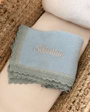 A personalized light blue knit baby blanket with a ruffled edge and the name 'Sebastian' embroidered in gold, displayed on a cream cushion inside a woven wicker basket.