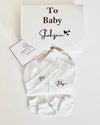 Personalized white baby gift set including an overall, socks, and a hat, presented in a gift box with a personalized message.