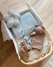 Baby gift set with blue knitted blanket, bear toy, and booties in a box on a wooden surface.
