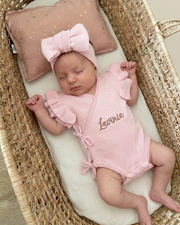 Newborn baby in a pink outfit with 'Lavinia' embroidered, lying in a wicker crib with a pink pillow.