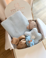 Collection of baby toys including a knitted blanket, teddy bear, star-shaped toy, and socks in a box with a brand logo.
