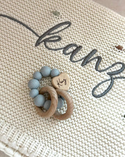 Teething ring with blue beads and a wooden heart on a textured surface with 'kean' embroidery.
