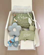 Gift box with baby items including a blanket, toys, and a plush flower on a beige background.