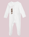 White baby cotton overall with personalized text and glitter details on the collar, displayed against a pink background.