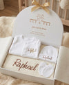 A personalized baby gift set in a white oval gift box with a gold ribbon, featuring a white long-sleeve baby shirt, matching hat, and bib with the name 'Raphael' embroidered in gold, displayed on a cozy beige blanket.