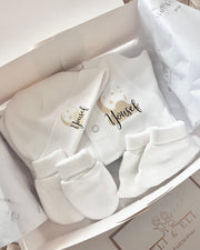 A set of beige baby gifts including a onesie, hat, socks, and mittens, presented in a box with a 'Welcome Baby' message.
