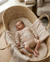 Newborn baby in a woven crib with a textured blanket and wooden rattle.
