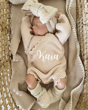 Newborn baby in a beige outfit with a matching hat, lying on a textured surface.