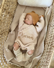 Newborn baby in a beige outfit with 'Karam' embroidered, lying on a textured surface.