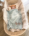 Newborn baby wrapped in a blanket with 'Agata' embroidered, lying in a woven basket.