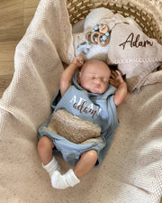 Newborn baby in a blue outfit with 'Adam' printed on it, lying in a basket with beige blanket and wooden toy.
