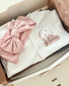 White cotton baby overall with a cloud pink bow, presented in an open gift box with the name 'Rania' printed on the front.