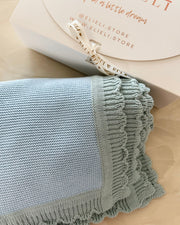A light blue knitted throw blanket with a ruffled edge, folded next to a white gift box with a cream ribbon. The box lid shows the brand name 'ELI' and website 'ELIELI.