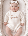 Baby in a white onesie with 'Georgina' embroidered, wearing a matching headband, lying in a textured basket.